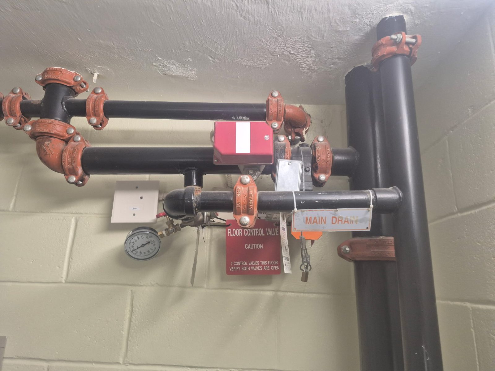Sprinkler floor control valve assembly — grooved black pipe, main drain, gauge and tamper switch in a riser room